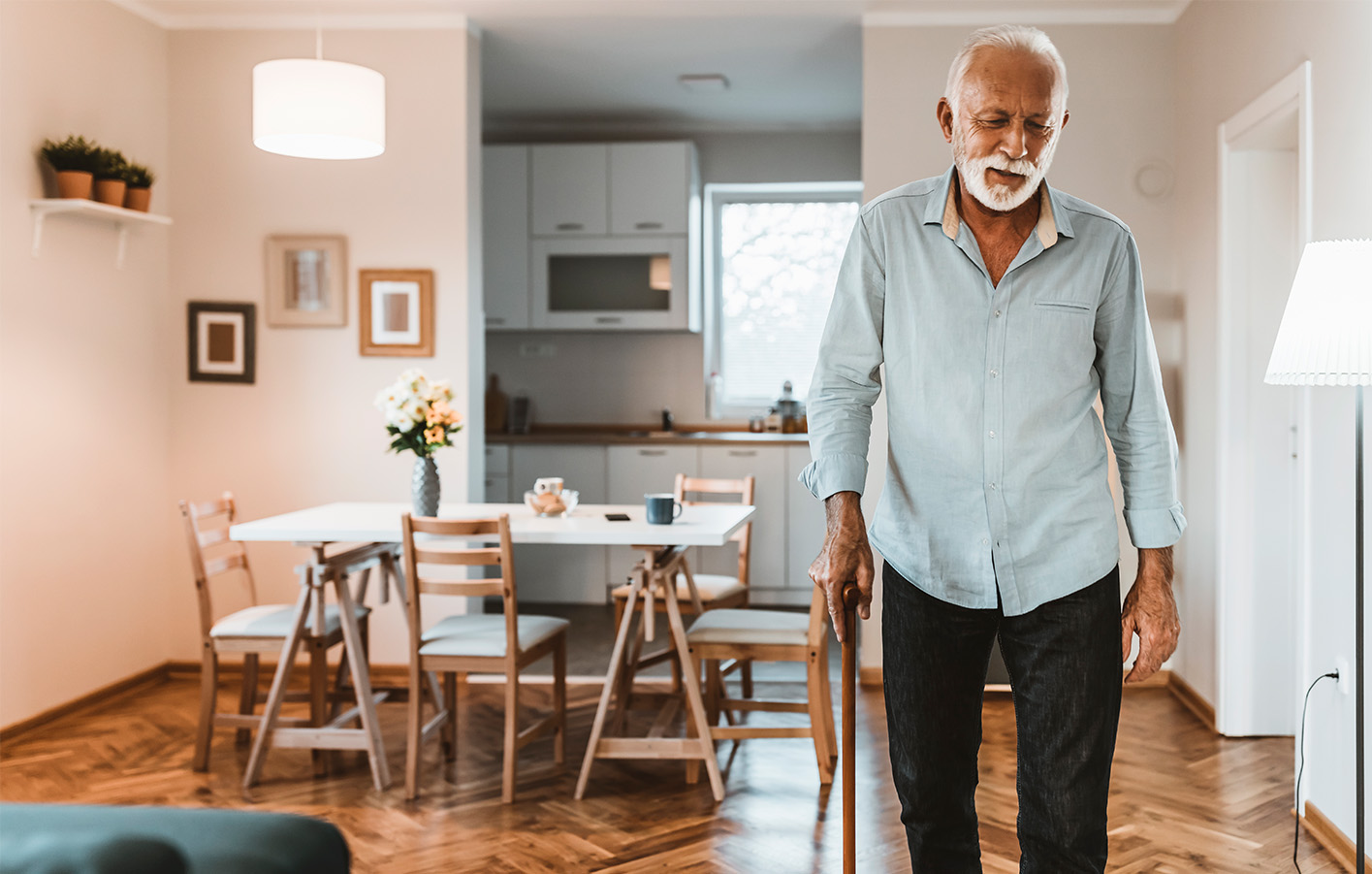 Älterer Herr mit weißem Haar und Krückstock geht vorsichtig durch ein helles Wohnzimmer mit Essbereich.